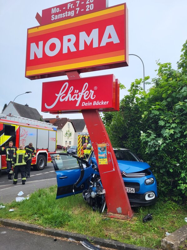 Verunfalltes Fahrzeug an einem Werbeschild eines Supermarktes, im Hintergrund Einsatzkräfte der Feuerwehr.
