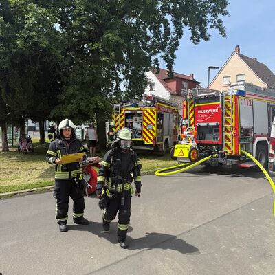 Bild vergr&ouml;&szlig;ern: Zwei Feuerwehreinsatzkräfte in Schutzausrüstung, dahinter zwei Löschfahrzeuge