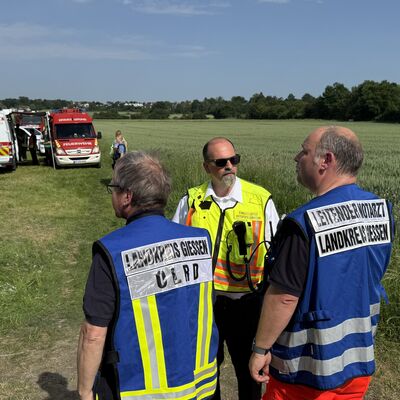 Bild vergr&ouml;&szlig;ern: Drei Einsatzkräfte auf einer Wiese vor Einsatzfahrzeugen
