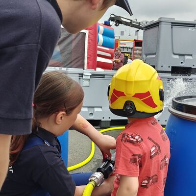 Bild vergr&ouml;&szlig;ern: Kind löscht mit Strahlrohr eine Feuerattrape und eine Jugendliche unterstützt. Ein anderer Jugendliche schaut zu.