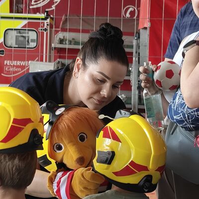 Bild vergr&ouml;&szlig;ern: Feuerwehrfrau trägt ein Kuscheltier und setzt einem Kind den Helm auf.