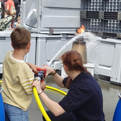 Bild vergr&ouml;&szlig;ern: Kind löscht mit Strahlrohr eine Feuerattrape und Feuerwehrfrau untersützt.