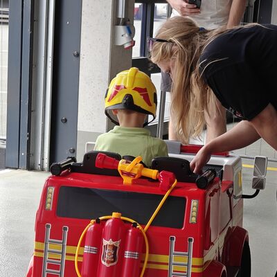 Bild vergr&ouml;&szlig;ern: Kind sitzt in Feuerwehrscooter und bekommt das Fahrzeug von einer Feuerwehrfrau erklärt.