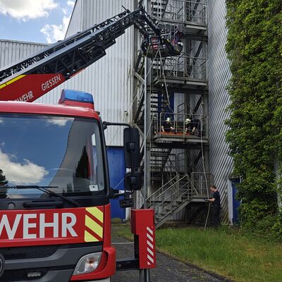Bild vergr&ouml;&szlig;ern: Im Vordergrund steht ein Drehleiterfahrzeug. Im Hintergrund sind zwei Einsatzkräfte auf einem Gerüst.