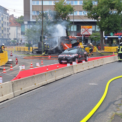 Bild vergr&ouml;&szlig;ern: Qualmender LKW vor einem Gebäude. Einsatzkräfte, die mit Löschmaßnahmen am LKW beschäftigt sind.