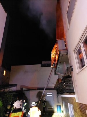 Einsatzkräfte vor einem Gebäude mit einem brenneden Balkon im 1. Obergeschoss. 