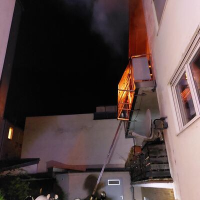 Bild vergr&ouml;&szlig;ern: Einsatzkräfte vor einem Gebäude mit einem brenneden Balkon im 1. Obergeschoss.
