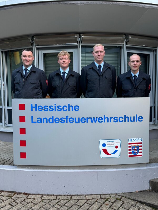 Bild vergr&ouml;&szlig;ern: Vier männliche Feuerwehreinsatzkräfte stehen hinter einem Schild der hessischen Landesfeuerwehrschule