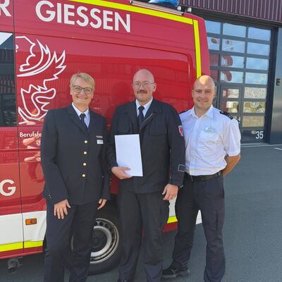 Bild vergr&ouml;&szlig;ern: Drei Feuerwehreinsatzkräfte in Uniform vor einem Einsatzfahrzeug.
