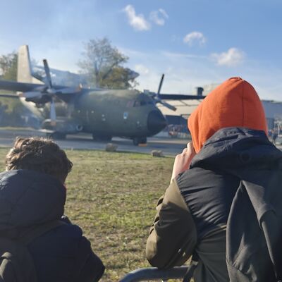Bild vergr&ouml;&szlig;ern: Drei Jugendliche beobachten ein Militärflugzeug im Hintergrund